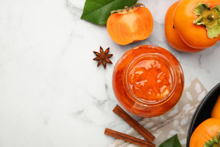 Jar of tasty persimmon jam and ingredients on white marble table, flat lay. Space for textの写真素材