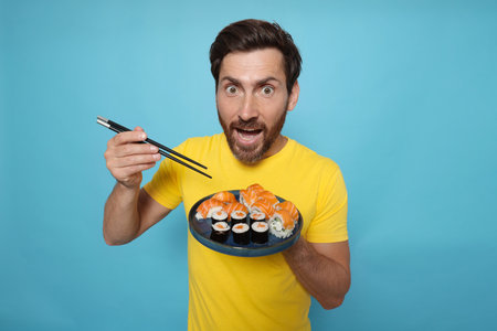 Emotional man holding plate with tasty sushi rolls and chopsticks on light blue backgroundの写真素材