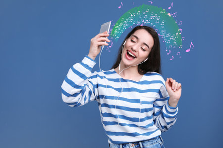 Happy woman listening to music through headphones on blue background, space for text. Music notes illustrations flowing from gadgetの写真素材