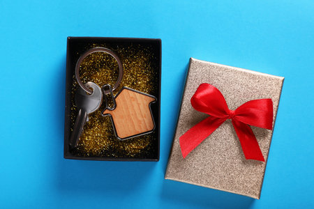 Key with trinket in shape of house and gift box on light blue background, flat lay. house warming...