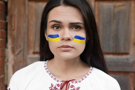 Young beautiful woman with drawings of Ukrainian flag on face near wooden doorの写真素材