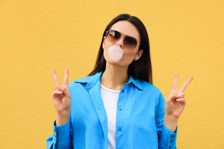 Beautiful young woman with sunglasses blowing chewing gum near orange wallの写真素材