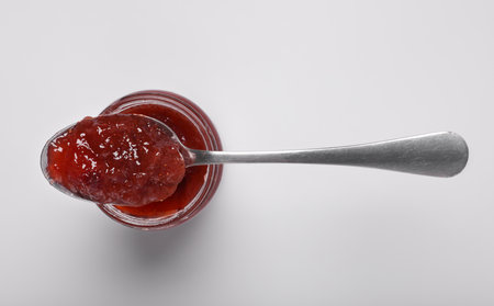 Spoon with tasty canned raspberry jam on jar against white background, top view