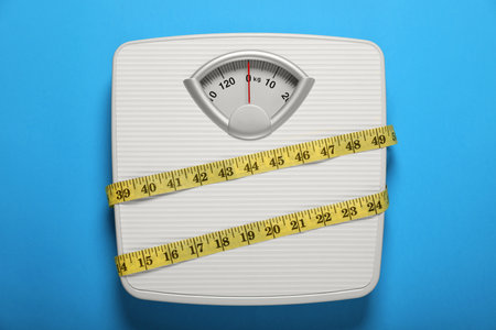 Bathroom scale tied with measure tape on light blue background, top view