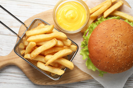 French fries in frying basket, tasty burger and sauce on white wooden table, flat lay