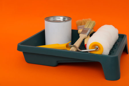 Can of orange paint, brushes, roller and container on color backgroundの写真素材