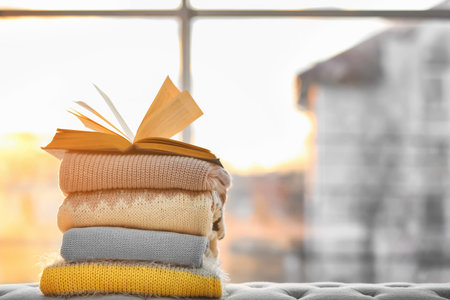 Stack of folded knitted clothes and open book against windowの写真素材