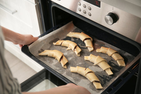Woman putting baking sheet with raw croissants in oven, closeup