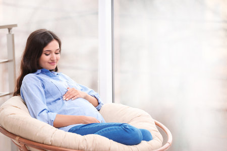 Beautiful pregnant woman sitting in lounge chair near windowの写真素材
