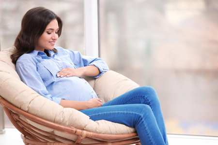 Beautiful pregnant woman sitting in lounge chair near windowの写真素材