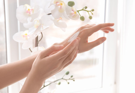 Young woman applying cream onto hands against windowの写真素材