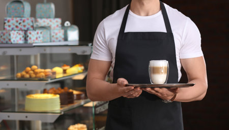 Young waiter holding tray with glass of coffee at workplaceの写真素材