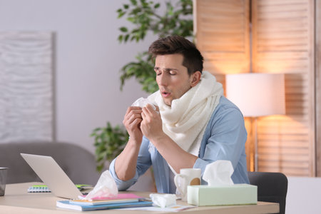Sad exhausted man with tissue suffering from cold while working with laptop at tableの写真素材