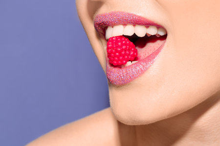 Beautiful young model with creative makeup holding candy in lips on color backgroundの写真素材