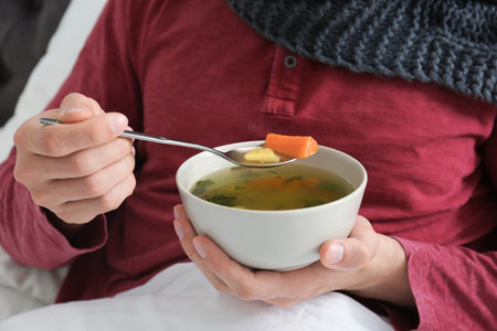 Sick young man eating broth to cure cold in bed at homeの写真素材
