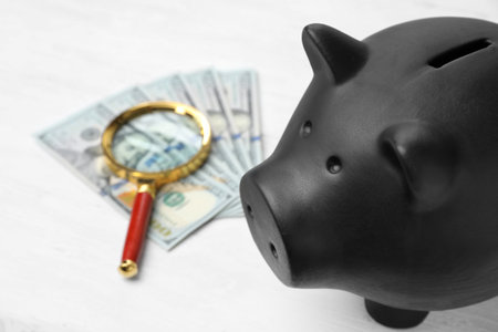 Black piggy bank, money and magnifying glass on tableの写真素材
