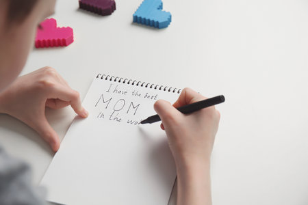Little child writing congratulations for Mother's Day in notebook on light backgroundの写真素材