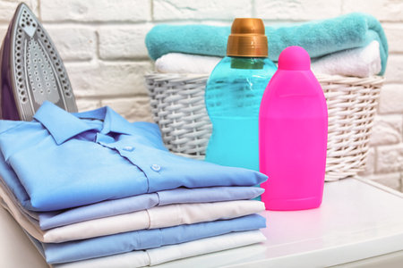 Stack of clean clothes, iron, detergents and basket with towels on tableの写真素材