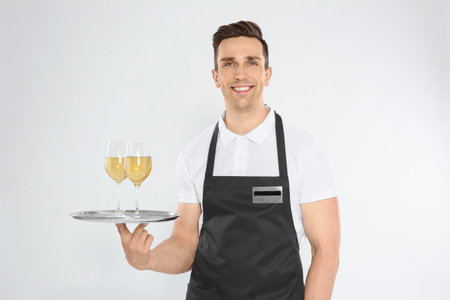 Handsome waiter holding tray with glasses of wine on light background