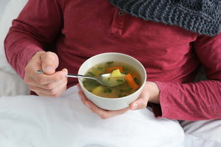 Sick young man eating broth to cure cold in bed at homeの写真素材