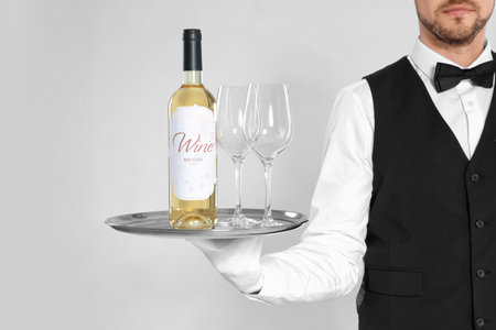 Young waiter holding tray with glasses and bottle of wine on light backgroundの写真素材