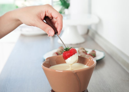 Woman dipping ripe strawberry into bowl with white chocolate fondue on tableの写真素材