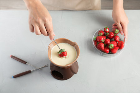 Woman dipping ripe strawberry into bowl with white chocolate fondue on tableの写真素材