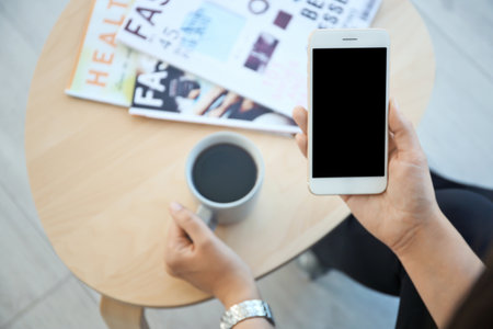 Young woman with cup of coffee holding mobile phone in hand at table indoorsの写真素材