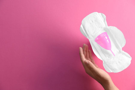 Woman holding hand near menstrual pad and cup on color background, top view. Gynecological careの写真素材