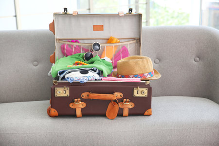 Open suitcase with different clothes and accessories for summer journey on sofa