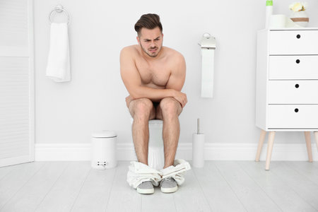 Young naked man suffering from diarrhea on toilet bowl at homeの写真素材
