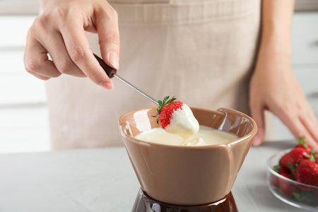 Woman dipping ripe strawberry into bowl with white chocolate fondue on tableの写真素材
