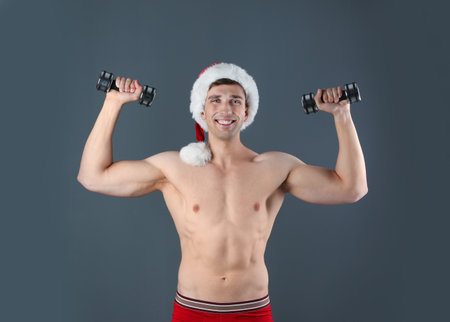 Young muscular man in Santa hat with dumbbells on gray backgroundの写真素材