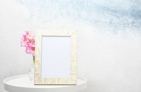 Blank frame and vase with flowers on table near light wall. Mock up design