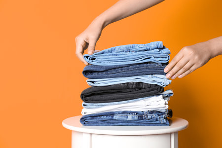 Woman arranging stack of different jeans on table against color background