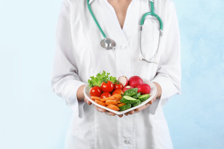 Female doctor holding plate with fresh vegetables on light background. Cardiac dietの写真素材