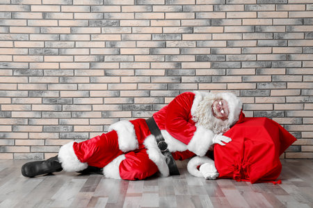 Authentic Santa Claus with big red bag full of gifts lying on floor near brick wallの写真素材