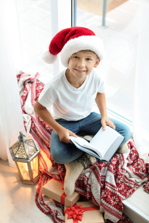 Cute little child in Santa hat reading Christmas story while sitting on windowsill at homeの写真素材