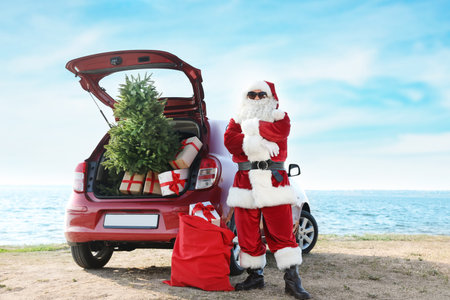 Authentic Santa Claus near red car with gift boxes and Christmas tree on beachの写真素材
