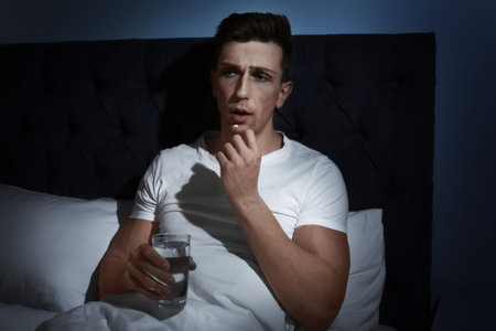 Young man with terrible headache taking pills in bed at nightの写真素材