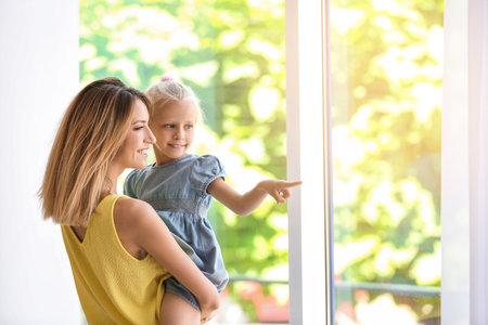 Young woman with cute little girl near window at homeの写真素材