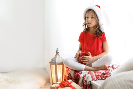 Cute little child in Santa hat with cup of cocoa sitting on windowsill at home. christmas celebrationの写真素材
