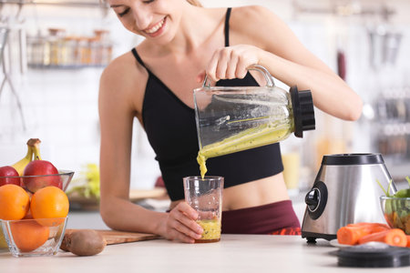 Young woman pouring tasty healthy smoothie into glass at table in kitchenの写真素材