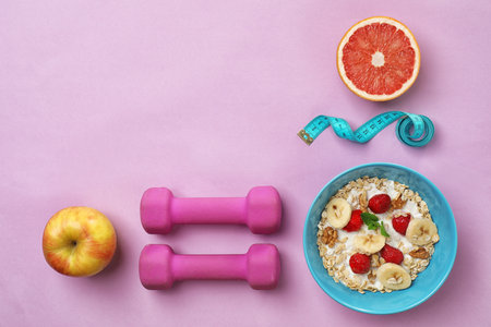Tasty oatmeal with fresh fruits in bowl, measuring tape and dumbbells on color background, top view....