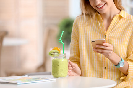 Young woman using mobile phone while drinking tasty healthy smoothie at table, indoorsの写真素材