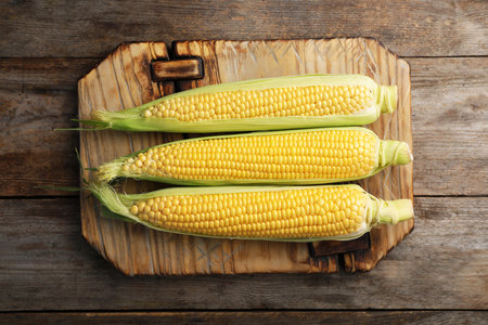 Tasty sweet corn cobs on wooden board, top view