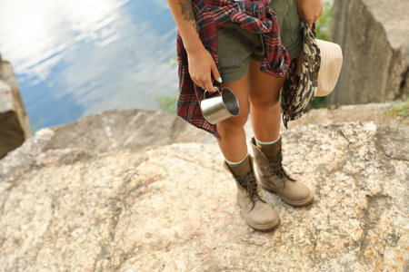 Young woman with hat and metal mug on rock, focus on legs. camping seasonの写真素材