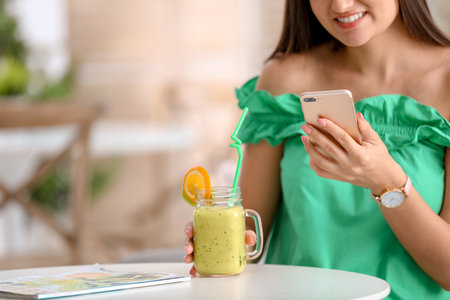 Young woman using mobile phone while drinking tasty healthy smoothie at table, indoorsの写真素材