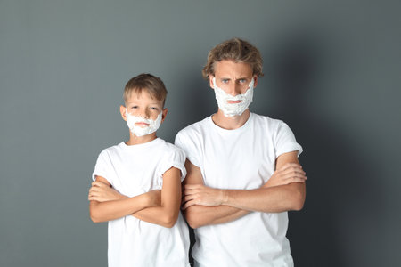 Father and son with shaving foam on faces against color backgroundの写真素材