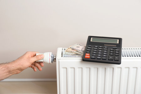 Man adjusting temperature of heating radiator with calculator and money on it indoorsの写真素材